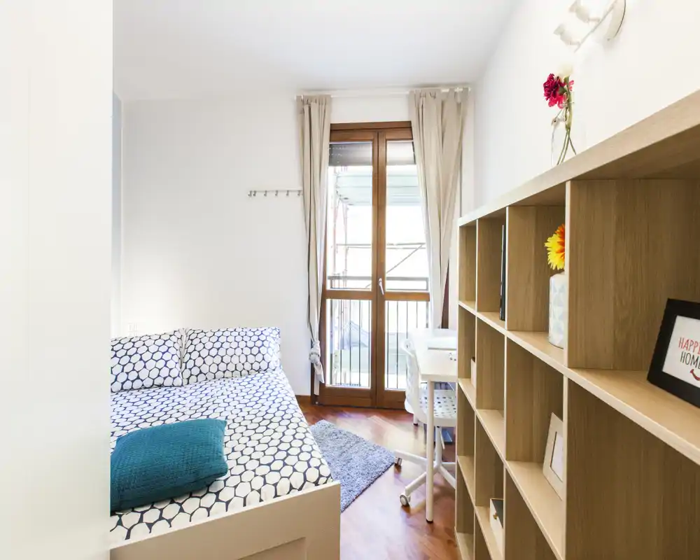 Camera da letto moderna e luminosa con letto matrimoniale con biancheria a motivo, libreria, piccolo angolo studio e porta balcone che illumina la stanza con luce naturale.