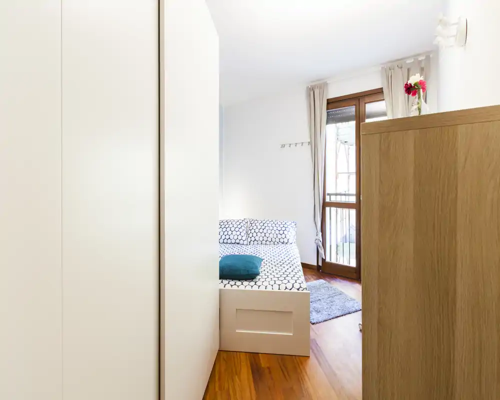 Camera da letto luminosa e moderna con biancheria a motivo, cuscino turchese e pavimento in legno; una grande finestra garantisce molta luce naturale e un tappeto caldo.