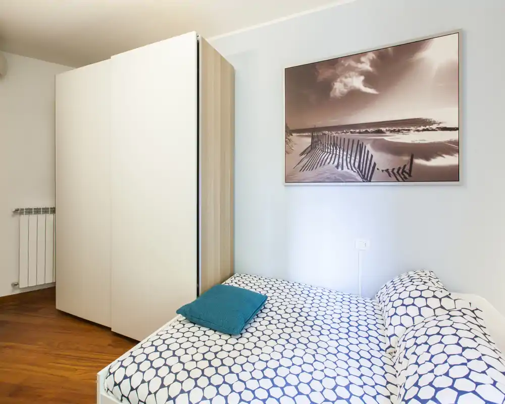 Camera da letto moderna e luminosa con letto matrimoniale a fantasia, cuscino turchese e ampio armadio; luce soffusa e quadro con tema marino creano un ambiente accogliente.