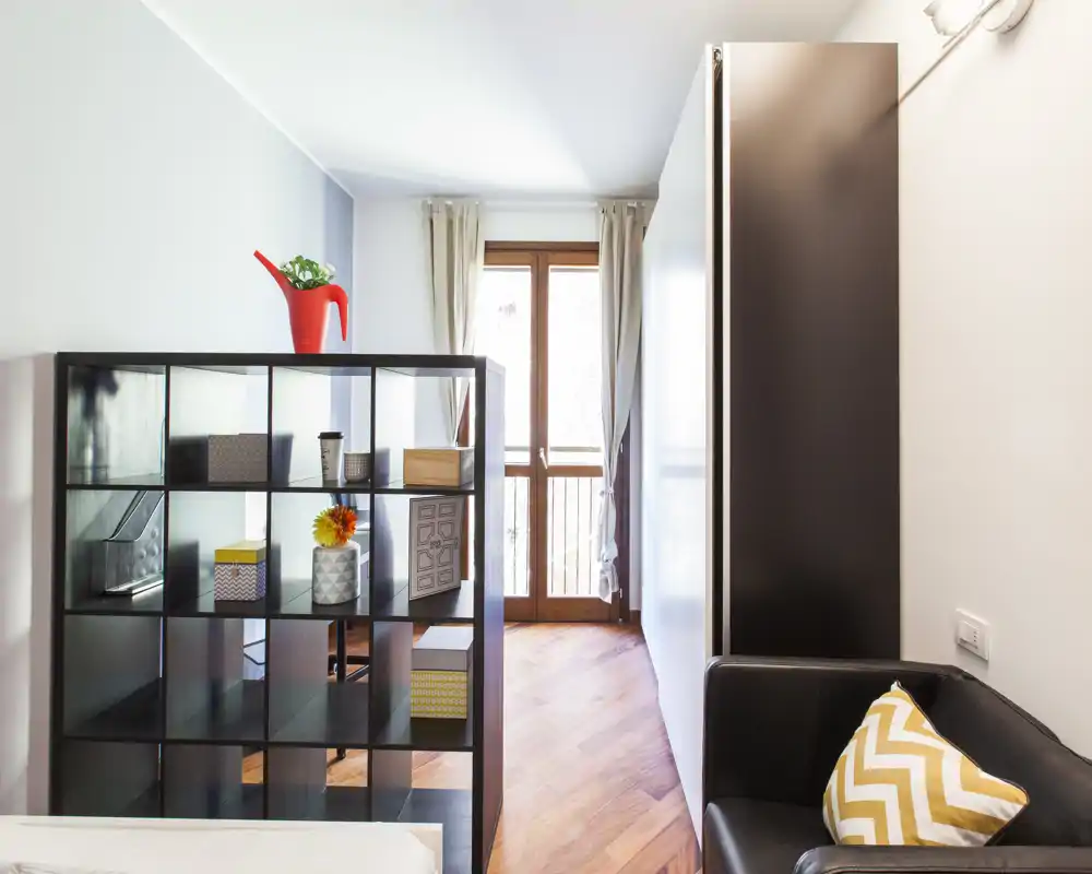 Bright, modern bedroom corner with a shelving unit acting as a room divider, wooden floor and a small seating area with patterned cushion.