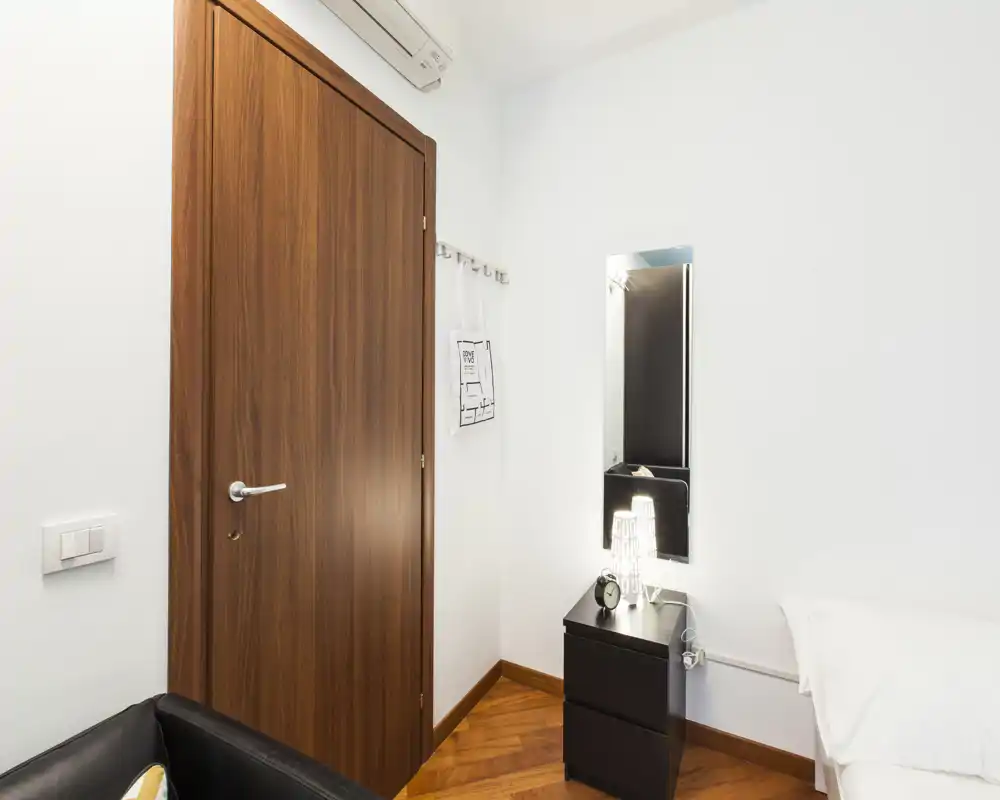 Small bedroom corner with a visible bedside table, lamp and a portion of the bed; clean white walls, wooden door and parquet floor create a modern, minimal look.