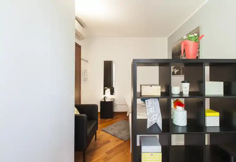 Bright, well-staged bedroom partially viewed through a room divider with a visible bed, bedside table, rug and comfortable armchair.