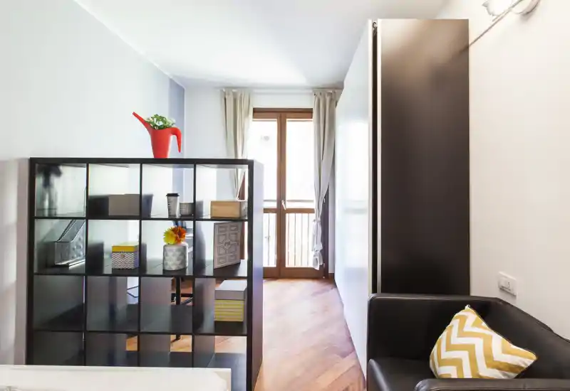Bright, modern bedroom corner with a shelving unit acting as a room divider, wooden floor and a small seating area with patterned cushion.