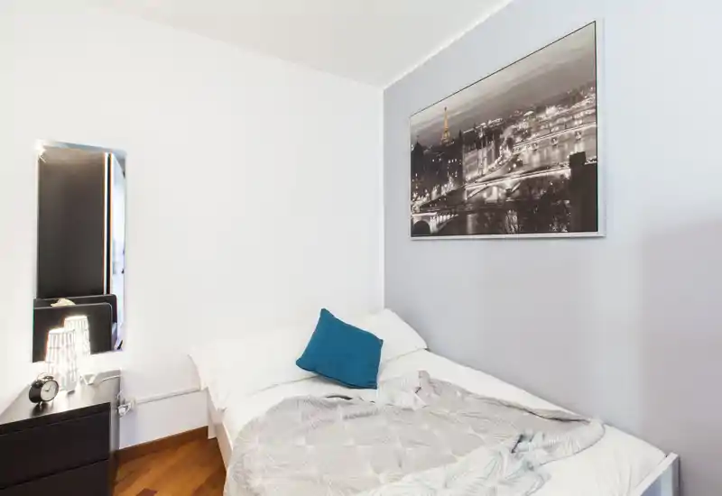 Bright, compact bedroom with a comfortable double bed, white linens and a teal accent cushion. Wall art and a mirror add style while a bedside cabinet with lamp provides practical storage and lighting.