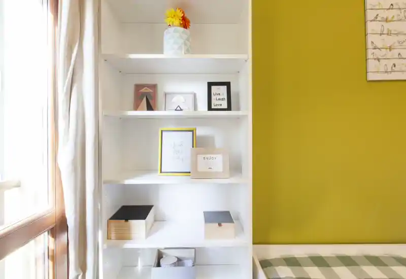Rincón de dormitorio luminoso con una estantería blanca, marcos decorativos y una cama a cuadros parcialmente visible junto a una ventana soleada.
