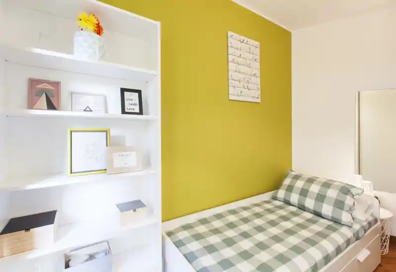 Habitación individual compacta y luminosa con edredón de cuadros, cojines decorativos y una pared de acento amarilla. Una estantería blanca y un espejo aportan almacenamiento y luz; ideal como estudio o habitación de invitados.