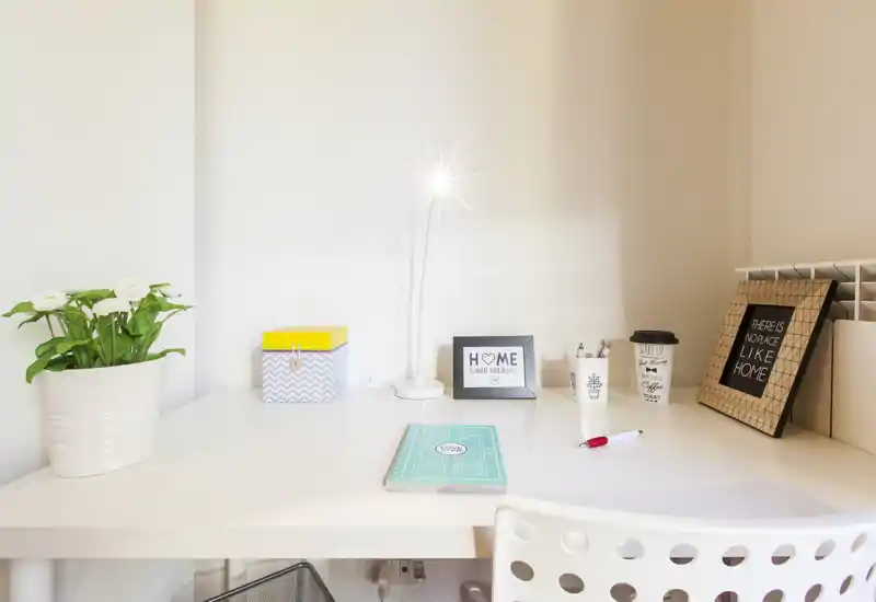 Bright, tidy workspace with a white desk, lamp, small plant and framed decor — a simple, welcoming home office/desk nook.