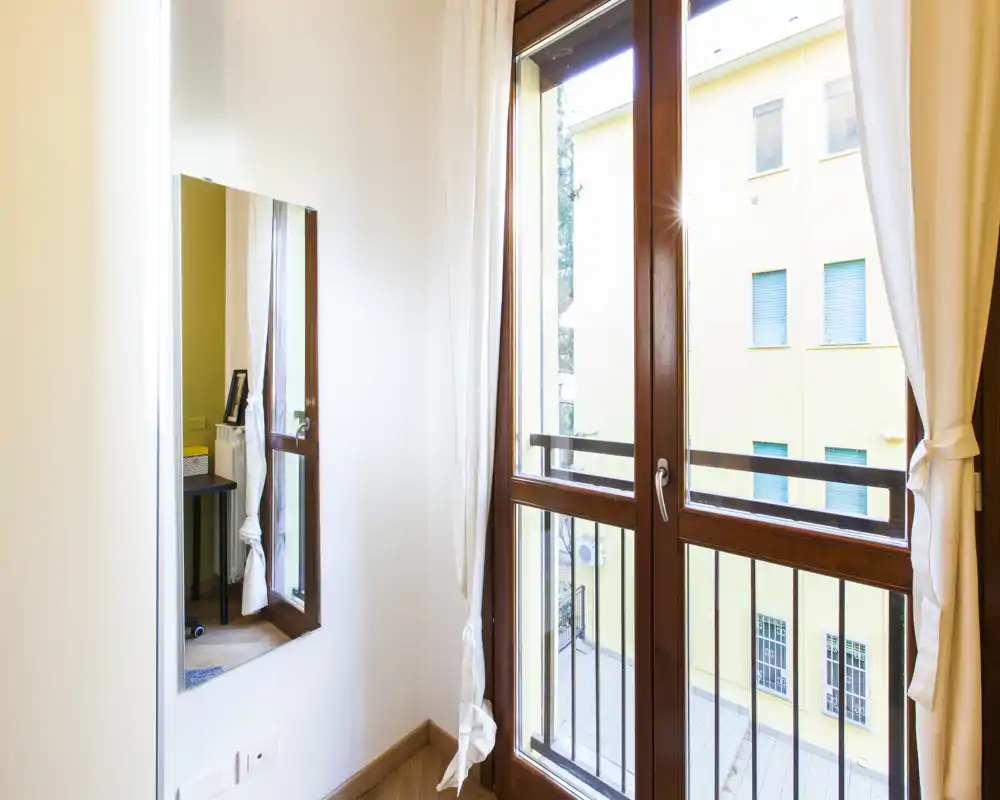 Vista su un piccolo balcone attraverso una porta finestra con cornice in legno che lascia entrare molta luce naturale; visibili tende leggere e un semplice parapetto.