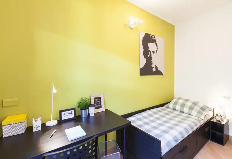 Camera singola luminosa e accogliente con scrivania ordinata e biancheria a quadretti. La parete gialla e l'opera d'arte decorativa conferiscono un tocco moderno e invitante, perfetto per uno studente o un ospite singolo.