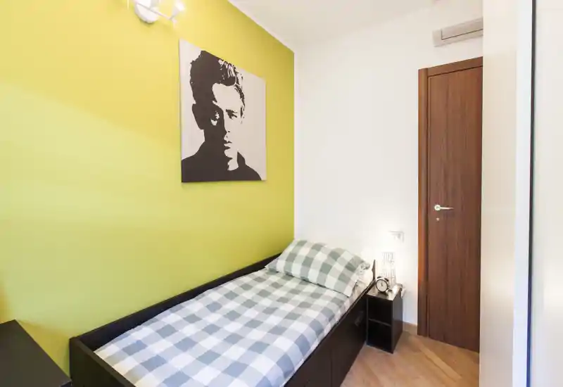 Camera singola compatta con letto a scacchi ordinato, pavimento in legno caldo e una parete accentata gialla con un quadro moderno. La stanza è luminosa, ordinata e arredata per un soggiorno confortevole.