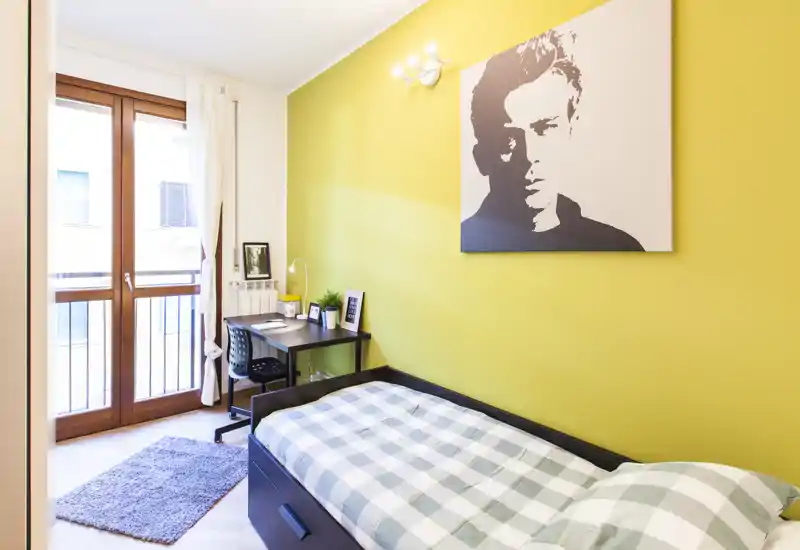Camera singola luminosa con letto comodo, scrivania e grande finestra sul balcone. La parete gialla e l’opera d’arte moderna creano un’atmosfera fresca e accogliente, ideale per studenti o professionisti.