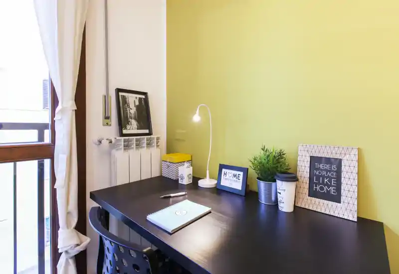 Angolo studio ben illuminato in zona giorno con scrivania in legno scuro, lampada da tavolo, stampe decorative e una piccola pianta: uno spazio di lavoro accogliente vicino alla finestra.