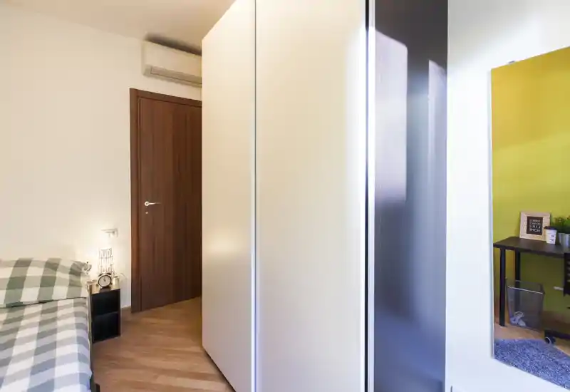 Camera compatta con angolo letto a quadri, comodino e lampada dal tono caldo; armadio moderno e uno specchio che riflette un'area lavoro adiacente.