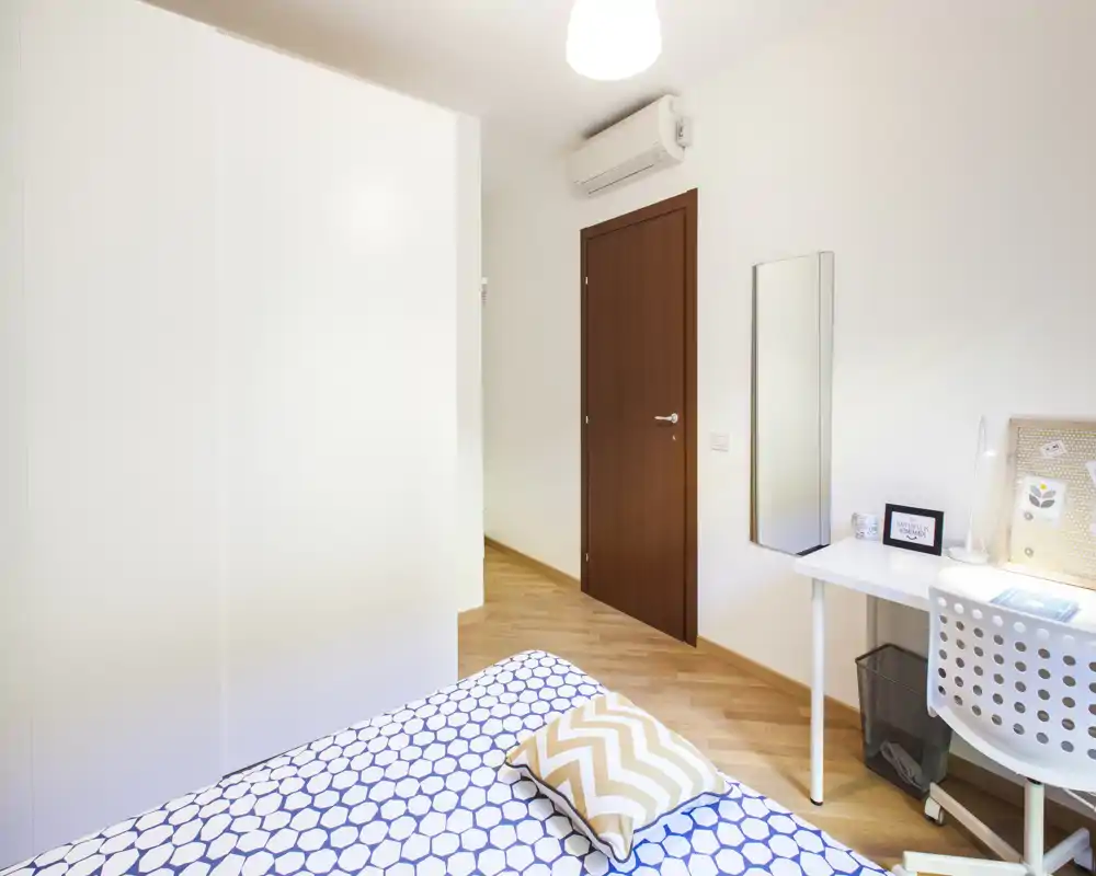 Dormitorio moderno y luminoso con colcha estampada, cojín decorativo, armario empotrado a la izquierda y un pequeño escritorio con silla y espejo junto a la puerta.