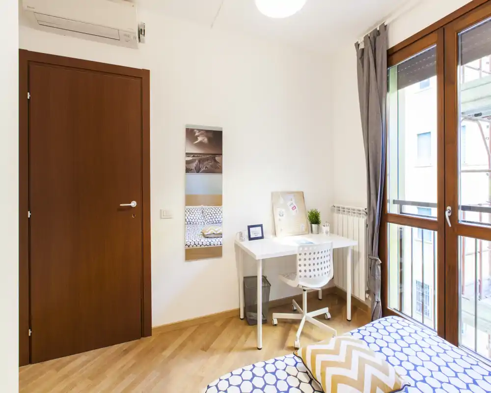 Rincón de dormitorio moderno y luminoso con cama estampada, pequeño escritorio y gran ventana que aporta mucha luz natural.
