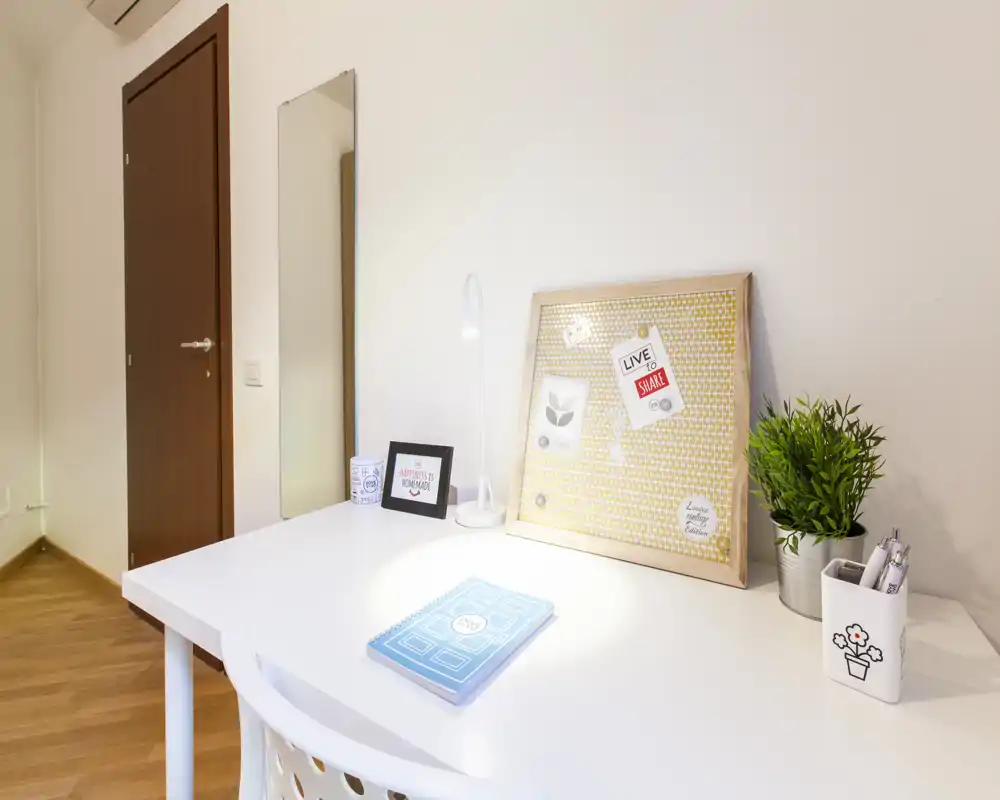 Rincón luminoso y ordenado con escritorio blanco, lámpara de trabajo, corcho y planta pequeña — ideal para estudiar o trabajar desde casa.