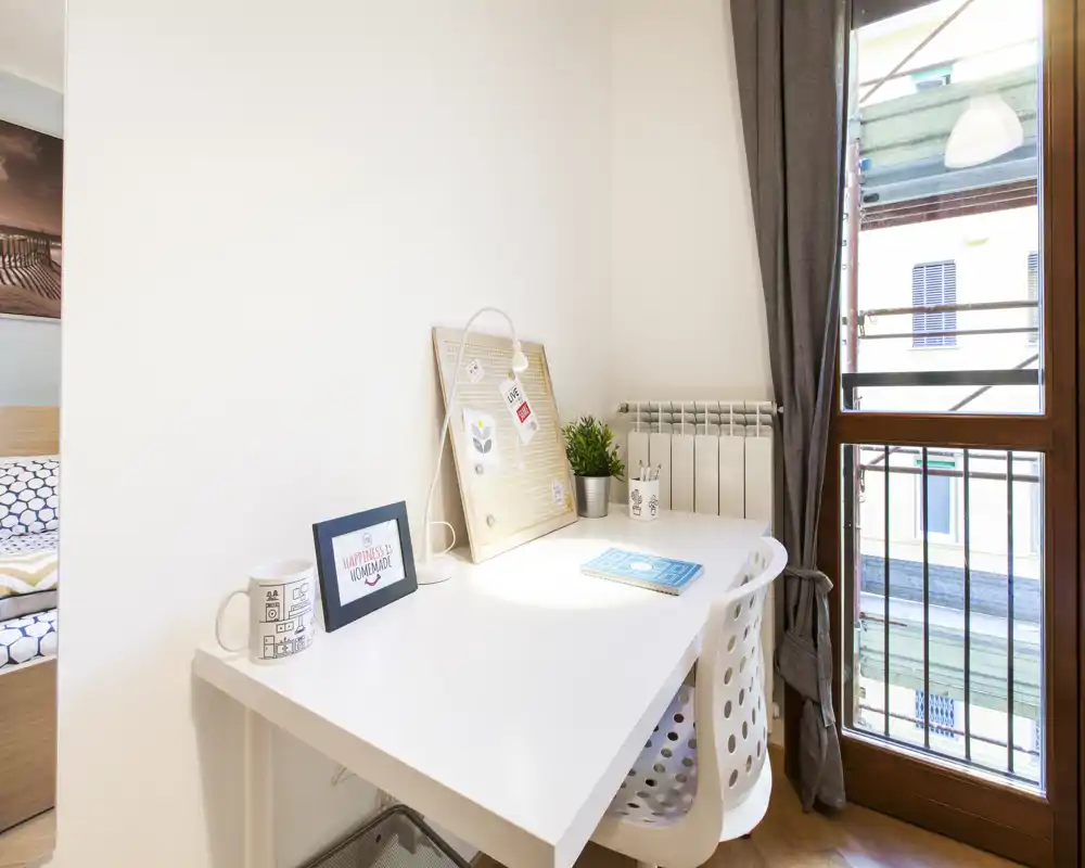 Rincón de dormitorio luminoso con un escritorio blanco junto a la ventana, tablero decorativo, pequeña planta y un vistazo de la cama en el borde del encuadre.