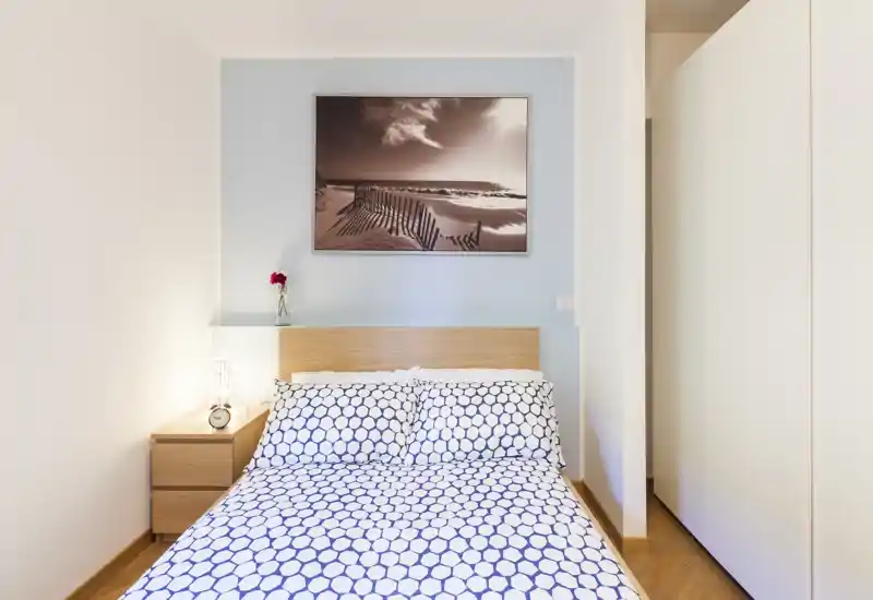 Camera da letto luminosa e dallo stile semplice con letto matrimoniale, biancheria a motivo, comodino con lampada e una stampa marina sopra la testiera.