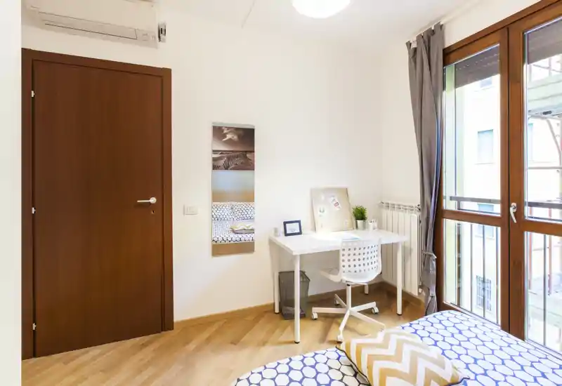 Angolo camera luminoso e moderno con letto a motif, piccolo scrittoio e ampia finestra che illumina la stanza.