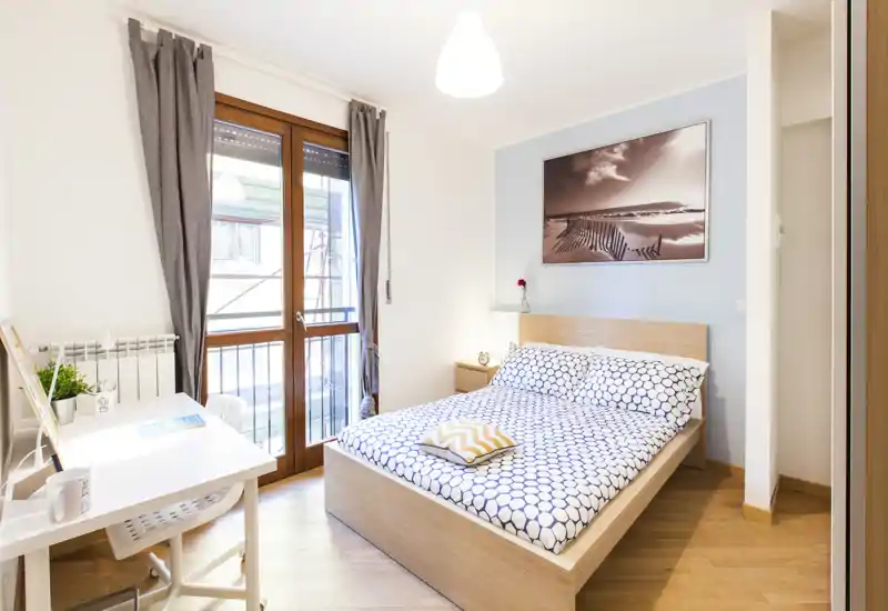 Camera da letto luminosa e moderna con letto matrimoniale, biancheria a motivo, comodino e scrivania vicino a una grande finestra che lascia entrare molta luce naturale.