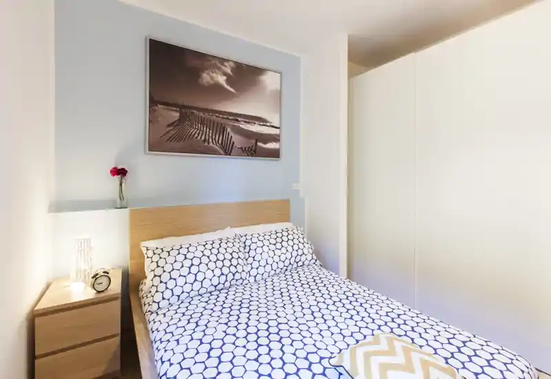 Camera da letto matrimoniale luminosa e ben arredata con biancheria a motivo, testiera in legno e guardaroba incassato. La stanza è curata con lampada da comodino e piccolo vaso decorativo.