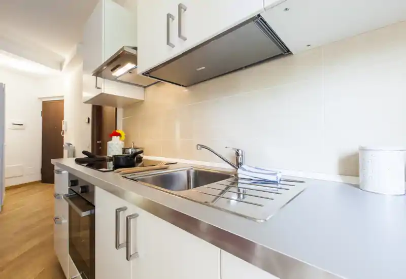 Modern compact kitchen with stainless-steel sink, induction hob and overhead extractor. Bright, clean countertop and light tiled backsplash ideal for a small apartment.