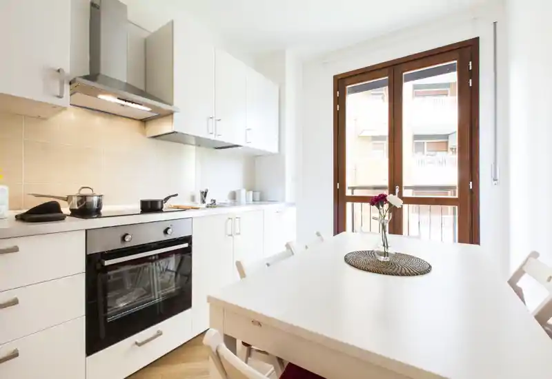 Bright modern kitchen with integrated oven, white cabinets and a dining table by a large window, styled with a simple vase of flowers.