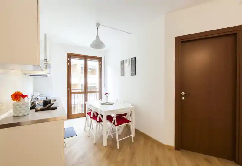 Bright compact kitchen with a small dining table next to a balcony door, white cabinets and warm wood flooring create a welcoming, functional space.