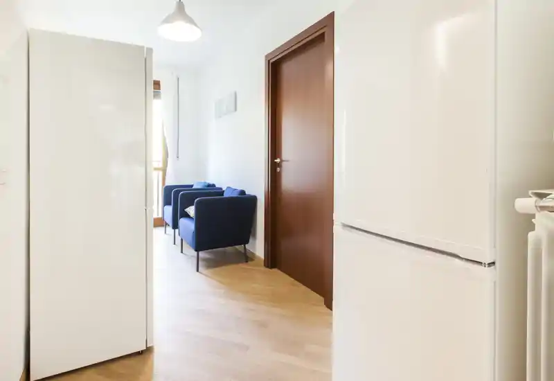 Small living area seen through a tight corridor: two blue armchairs by a window, warm wooden floor and soft natural light creating a welcoming nook.