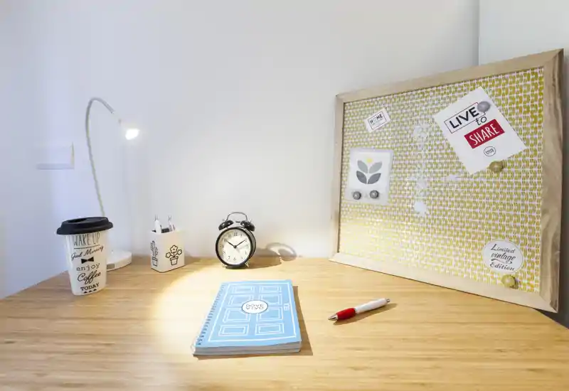 Coin bureau lumineux et bien rangé avec plateau en bois, lampe de bureau, réveil, carnet et un tableau d'affichage décoratif — parfait pour un espace de travail à la maison chaleureux.