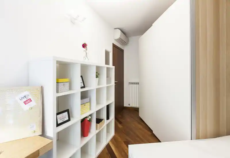 Compact bedroom corner with a visible mattress corner, white shelving unit and warm wooden floor; bright, clean and simply decorated.