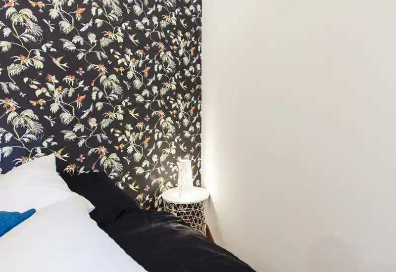 Coin de chambre lumineux et stylé avec papier peint à motifs, table de chevet et lampe ; une partie du lit avec oreillers et linge est visible.