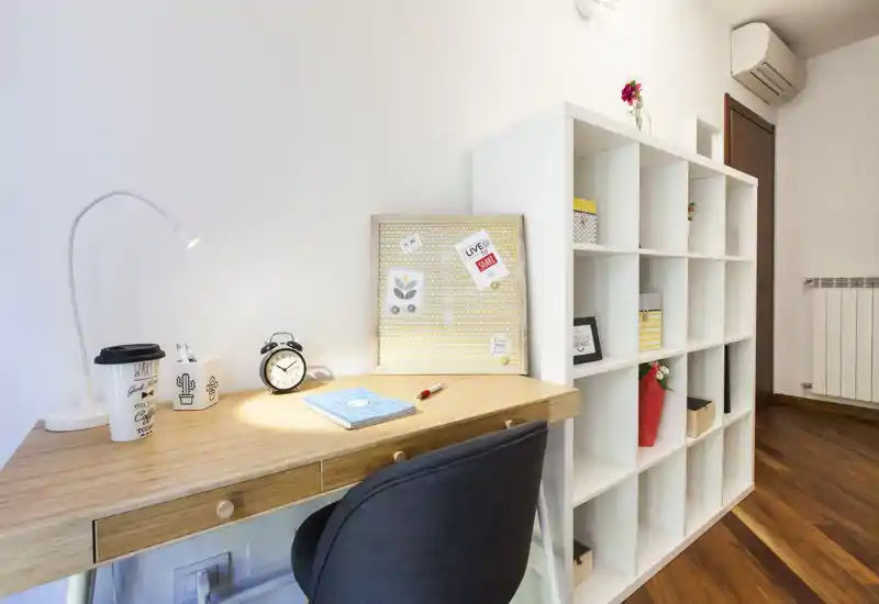 Coin de séjour lumineux et moderne aménagé en espace de travail à domicile avec bureau en bois, lampe, réveil et étagères ouvertes — un endroit chaleureux et bien éclairé, idéal pour le télétravail ou les études.