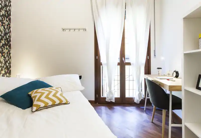Bright, modern bedroom with a neatly made bed, decorative cushions and a small desk by a large window letting in natural light.