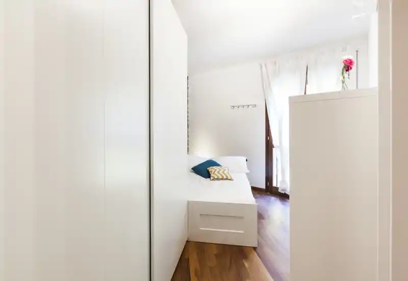 Bright, minimal bedroom with a neat white bed, decorative cushions and natural light from a balcony window; warm wooden floor and clean modern storage create a welcoming atmosphere.