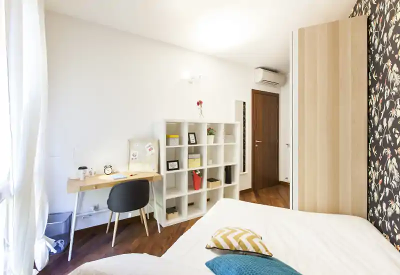 Bright, modern bedroom with a comfortable double bed, decorative cushions and a small workspace by the window. The room features warm wooden floors, a shelving unit for storage and stylish patterned wallpaper on one wall.