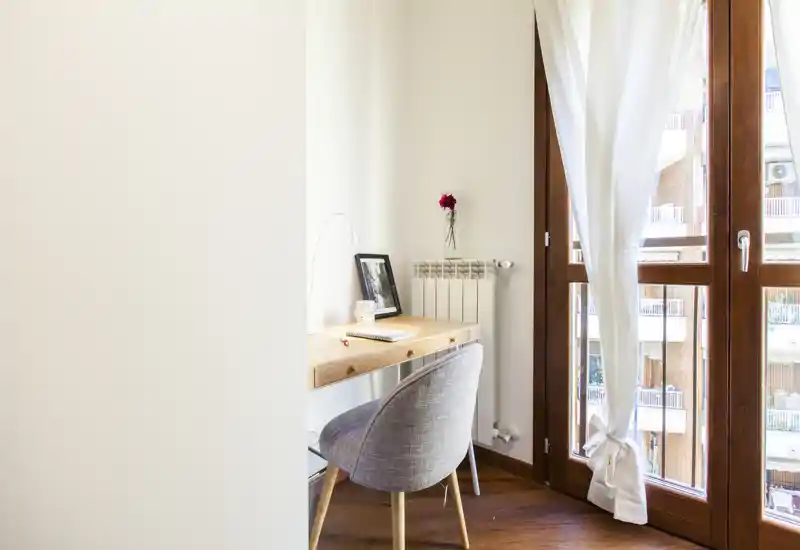 Angolo lavoro luminoso in soggiorno con scrivania in legno, sedia imbottita e ampia finestra che lascia entrare luce naturale.