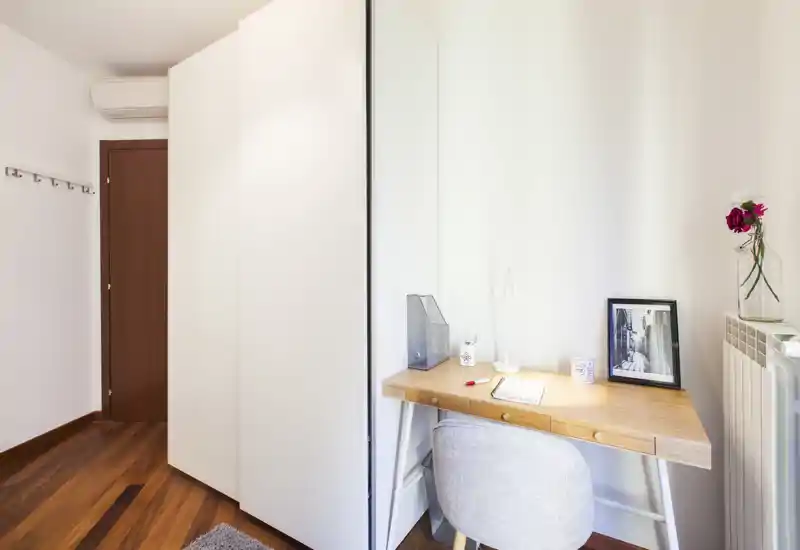 Rincón de dormitorio compacto y luminoso con escritorio de madera y silla tapizada junto a un armario blanco; detalles decorativos cuidados como foto enmarcada y flores.