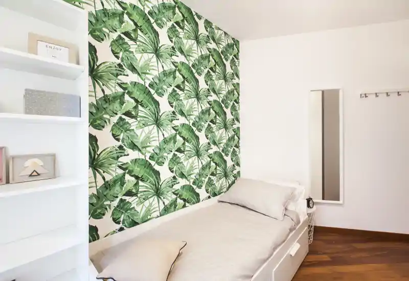 Dormitorio pequeño y con estilo, con cama individual frente a una pared decorativa botánica verde. Ropa de cama limpia, espejo y estanterías que hacen el espacio luminoso y acogedor — ideal para una estancia cómoda.