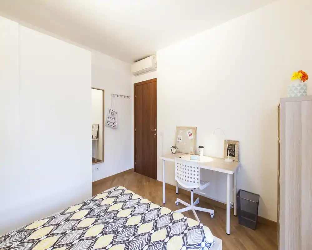 Dormitorio moderno y luminoso con cama con estampado, escritorio sencillo y silla junto a la pared — ordenado, bien iluminado y acogedor.