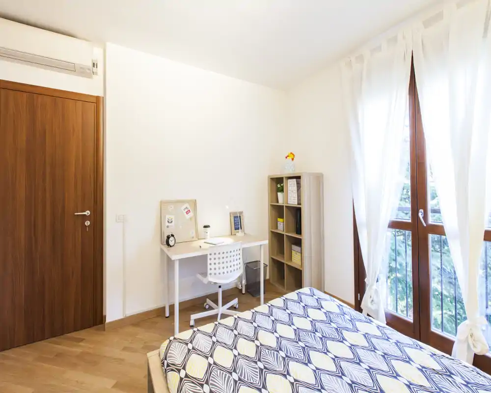 Dormitorio luminoso y bien presentado con cama doble estampada, escritorio y estantería junto a una gran ventana que aporta luz natural.