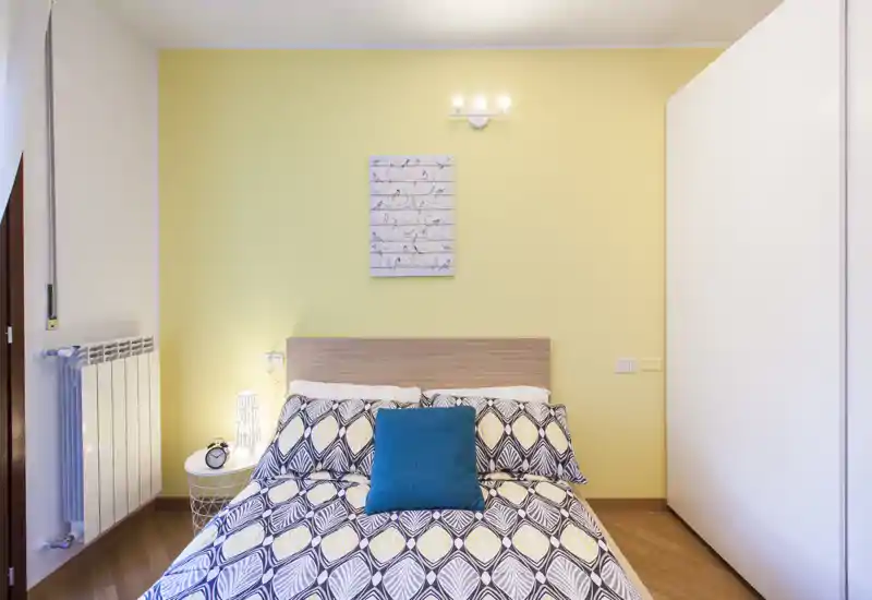 Dormitorio luminoso y ordenado con ropa de cama estampada, cojín turquesa y cabecero de madera frente a una pared amarillo suave. Mesita, lámpara y armario completan un ambiente acogedor, ideal para anuncios.