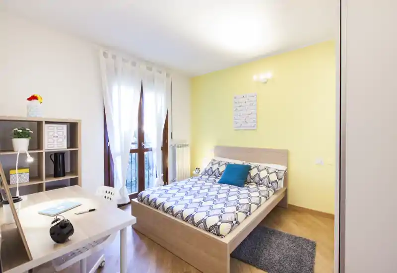 Dormitorio luminoso y bien decorado con cama doble, ropa de cama con estampado y cojín. Luz natural entra por cortinas translúcidas sobre suelos de madera; dispone de zona de trabajo y estantería.