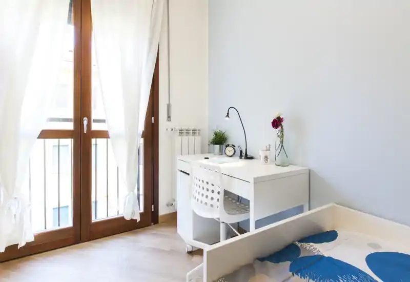 Rincón de dormitorio luminoso y ordenado con cama individual, escritorio blanco compacto y una gran ventana que aporta luz natural: ideal para trabajar y descansar.