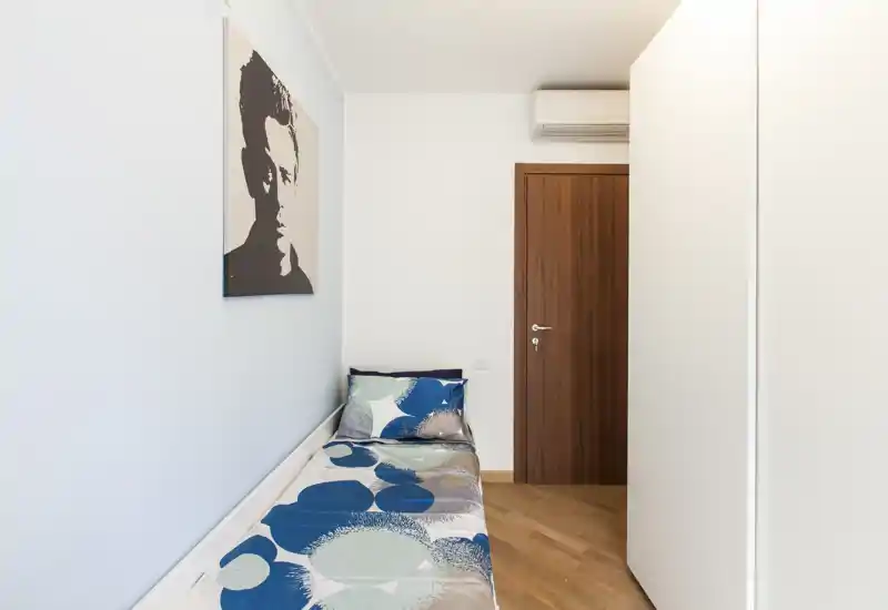 Habitación individual compacta con cama de una plaza estampada, paredes claras y suelo de madera cálida. Espacio luminoso y minimalista con cuadro en la pared y aire acondicionado sobre la puerta.