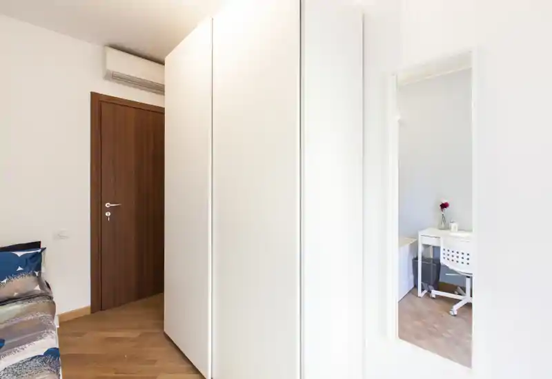 Rincón de dormitorio con cama parcialmente visible, amplio armario blanco y espejo de cuerpo entero que refleja una zona de escritorio; luminoso y limpio, apto para anuncios de alquiler.