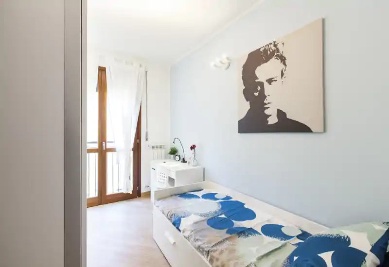 Dormitorio compacto y luminoso con cama individual, escritorio y luz natural desde la puerta al balcón — ideal para un estudiante o persona sola.