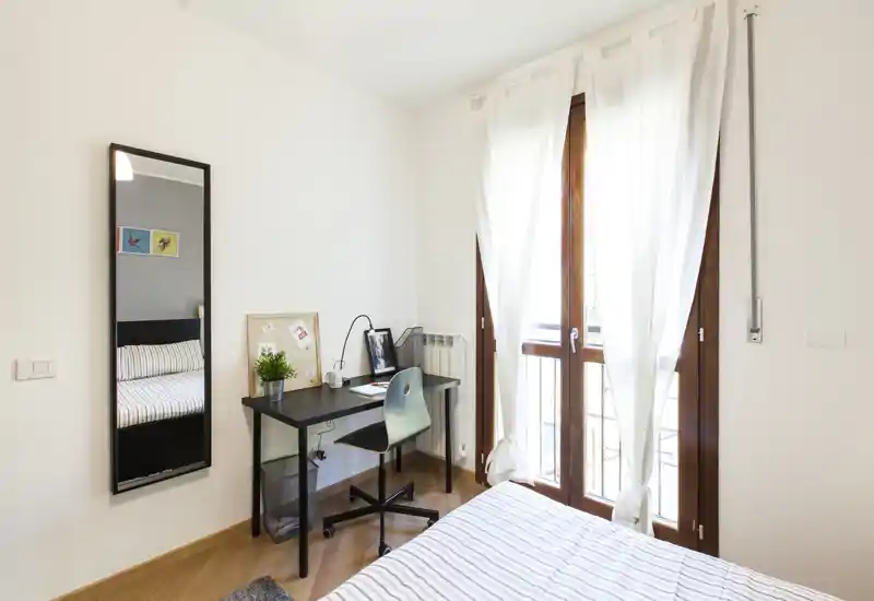 Camera da letto luminosa e ben sistemata con letto a righe, una scrivania compatta e sedia accanto a grandi finestre che lasciano entrare molta luce naturale.