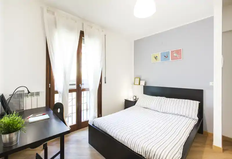 Camera da letto luminosa e ben arredata con letto matrimoniale, comodino e scrivania accanto a una grande finestra che lascia entrare molta luce naturale.