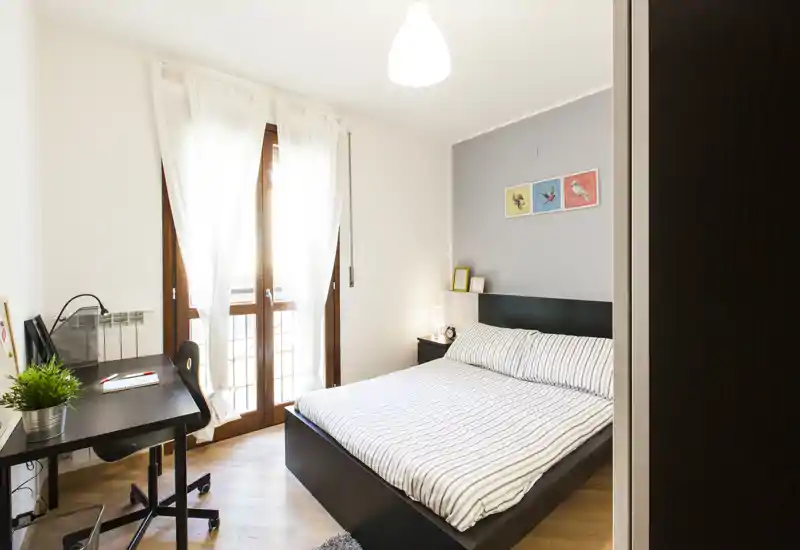 Camera da letto moderna e luminosa con letto matrimoniale, comodini e una scrivania vicino alla finestra. La luce naturale attraversa le tende leggere creando un'atmosfera calda e accogliente.